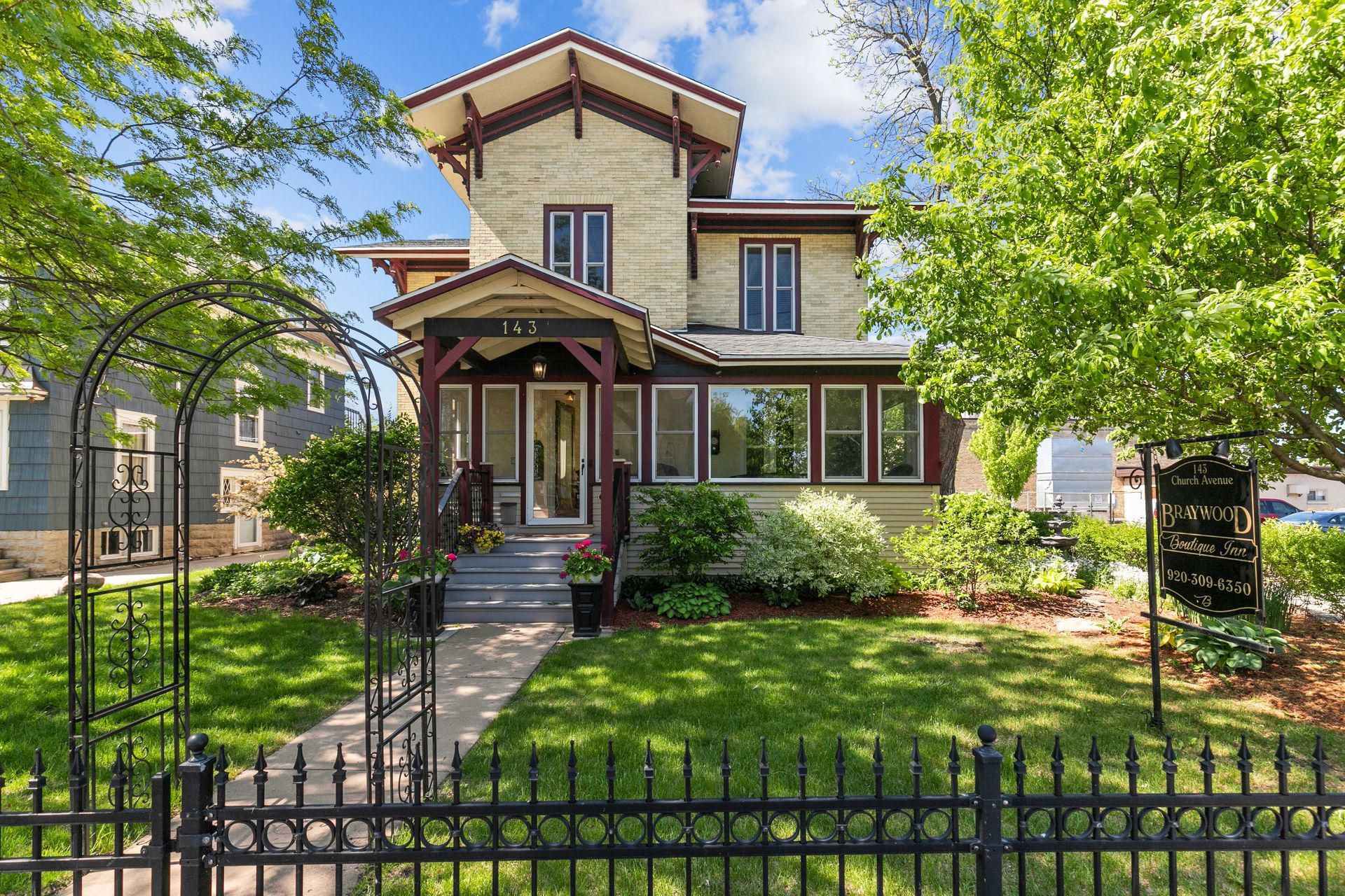 Two-story Victorian house with beige exterior, black fence, green lawn and trees.