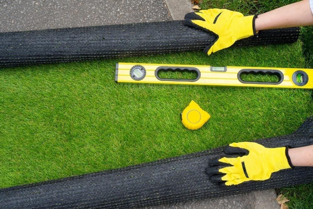 Hands unroll artificial grass next to measuring tools on a surface.