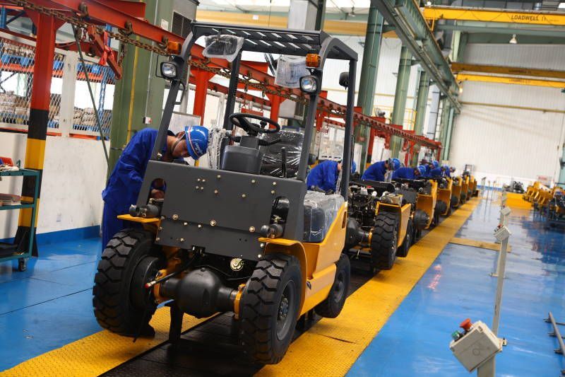 Forklifts on an assembly line, being worked on by people in blue coveralls inside a factory.