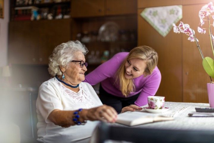 Woman leaning toward smiling seated elder at a table in a cozy kitchen, with a teacup and flowers nearby