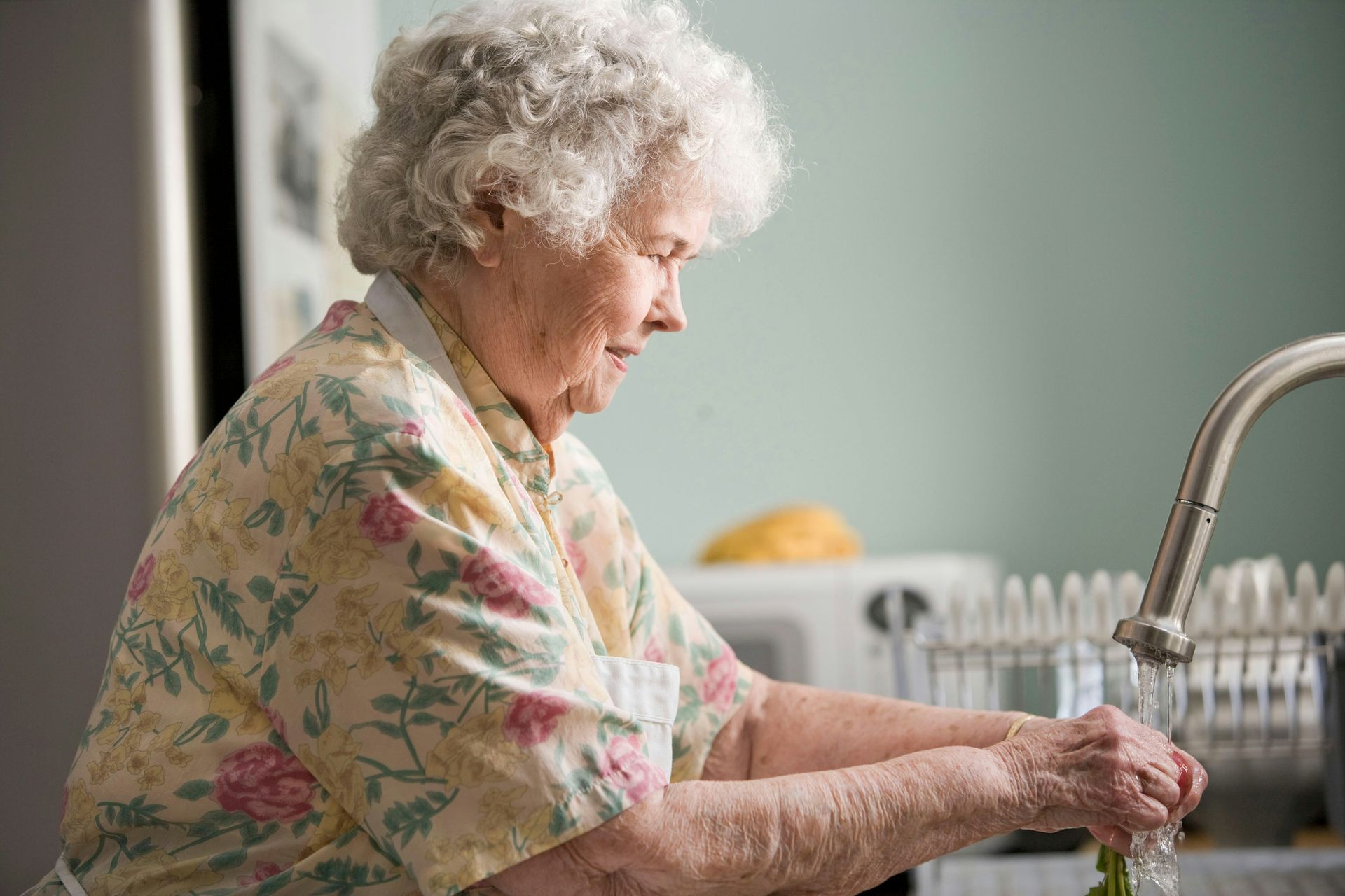 Elderly woman washing dishes at a kitchen sink, wearing a floral blouse.