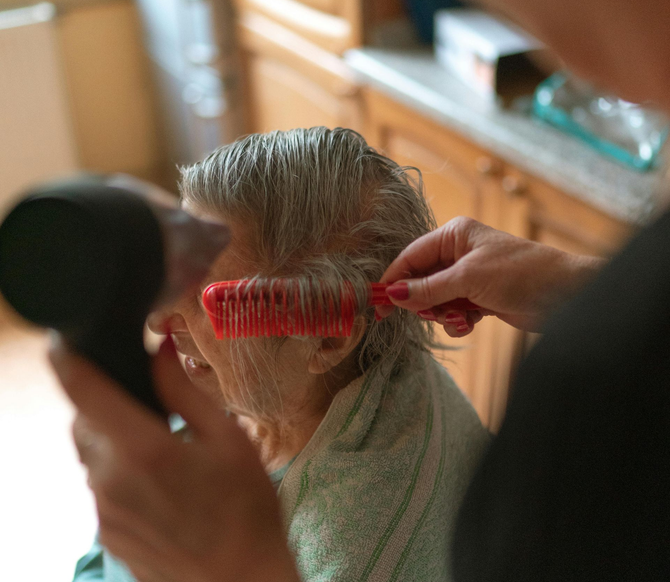 Hair being brushed with a red comb while someone holds a handheld mirror in a sunlit room