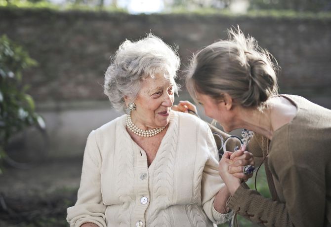 Two women smiling and talking closely outdoors, one in a cream sweater and the other in a green jacket
