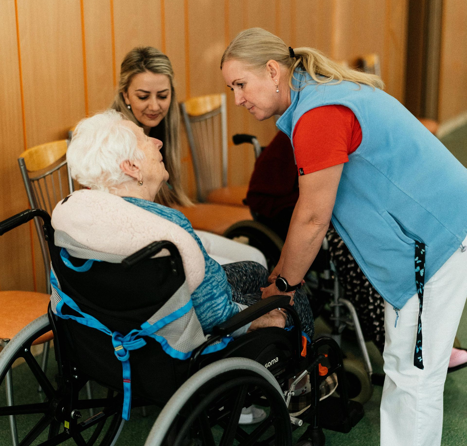 Caregiver helping an elderly woman in a wheelchair while another woman smiles nearby in a room