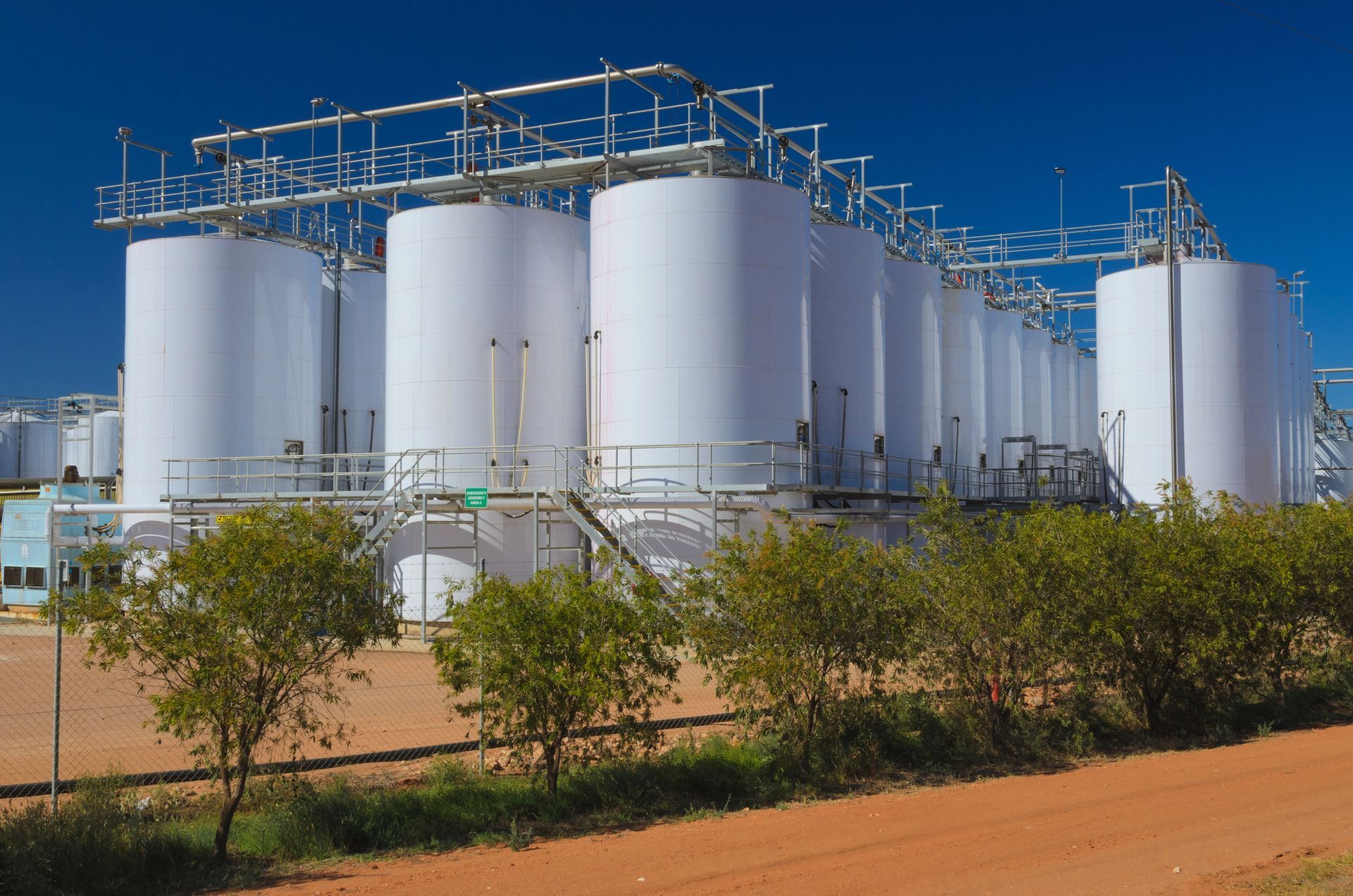 Serbatoi di stoccaggio bianchi in un ambiente industriale, tubazioni nella parte superiore, cielo azzurro sullo sfondo.