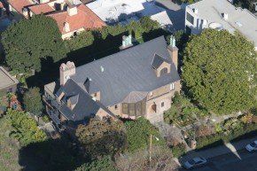 Aerial view of a large brownstone house with a dark gray roof, surrounded by trees and residential buildings.