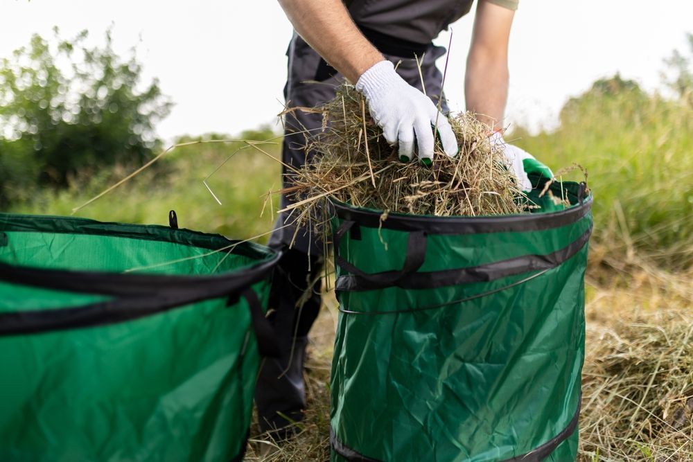Person Wearing Gloves Putting Dry Grass Into a Green Pop-up Yard Waste Bag — All Suburbs Garden Bags in Holloways Beach, QLD