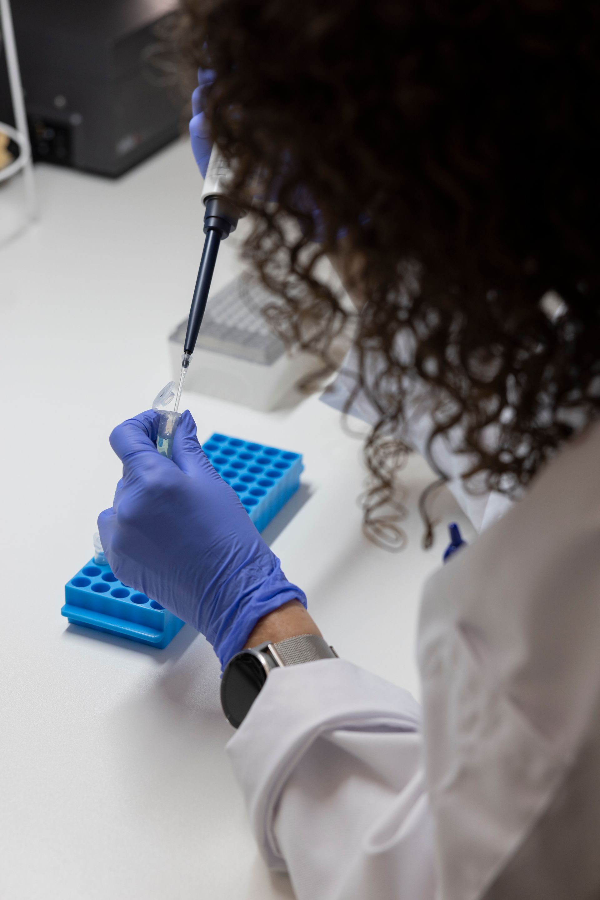 Cynthia Dirks zit met een  laboratoriumjas en blauwe handschoenen gebruikt een pipet om vloeistof over te brengen naar een klein reageerbuisje.