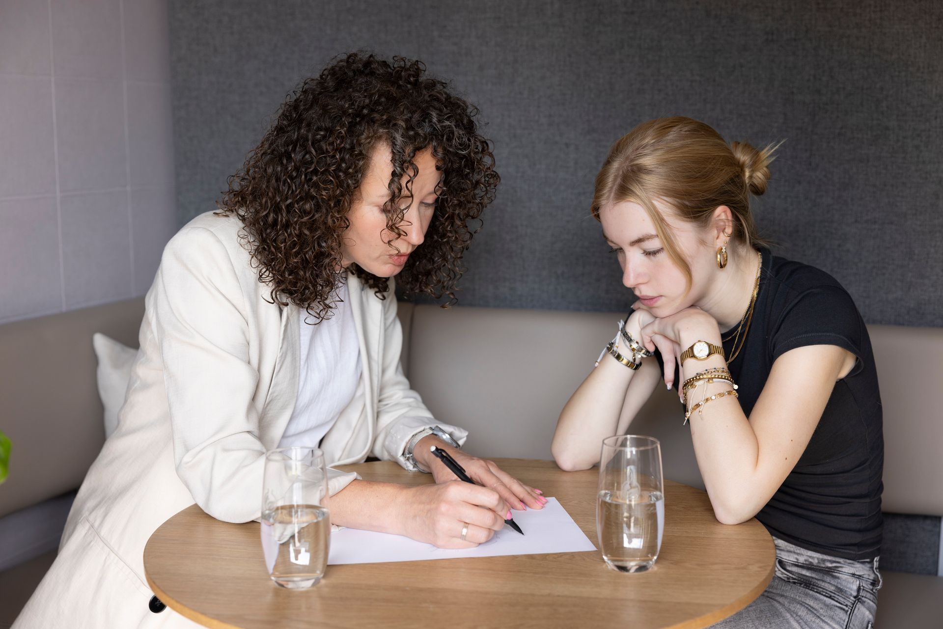 Twee mensen zitten aan een ronde tafel; de een schrijft op papier terwijl de ander toekijkt met de kin op de handen.