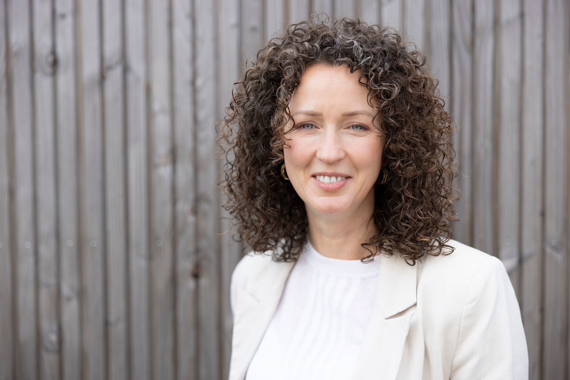 Metabool therapeut Cynthia Dirks lacht naar de camera, gekleed in een wit overhemd en blazer, tegen een achtergrond van houten latten.