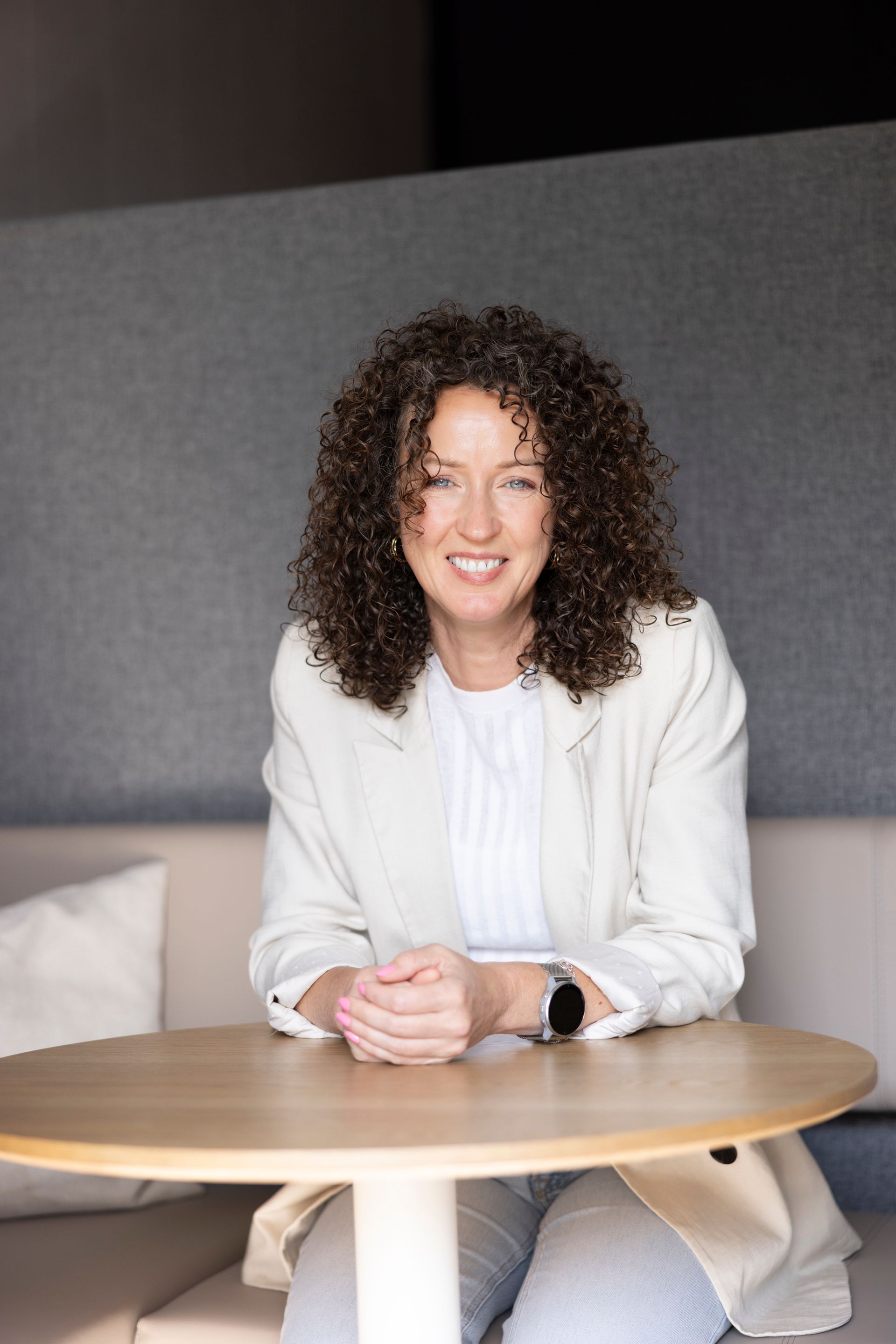 Metabool therapeut Cynthia Dirks gekleed in een witte blazer, zit aan een ronde tafel en glimlacht in een moderne kantooromgeving.