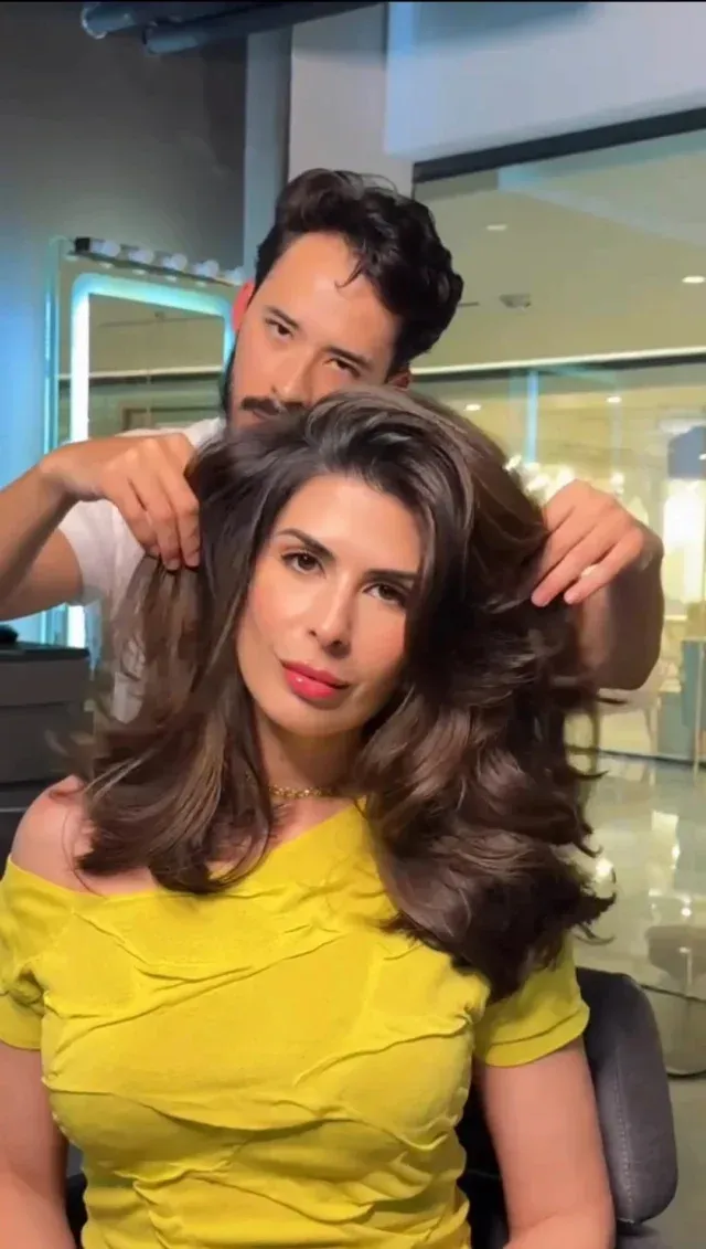 Woman in yellow top with stylist fluffing her brown hair in salon.