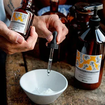 Person adding liquid from a brown bottle with a dropper into a white bowl. Another bottle and a pump bottle are visible.