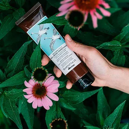 A hand holding a brown hair conditioner tube with birds on a blue background, surrounded by plants and flowers.
