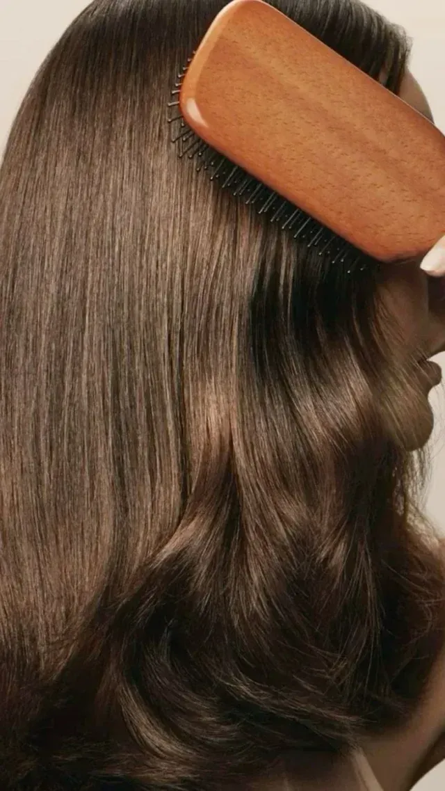 Person brushing long, dark brown hair with a wooden paddle brush.