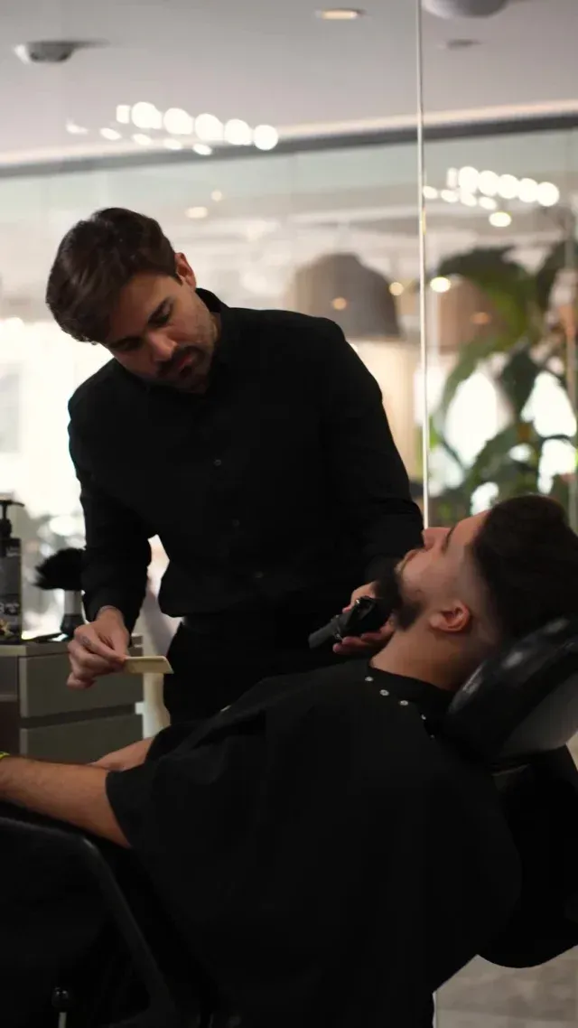 Barber shaves a client's beard in a modern salon. Both wear black. The client reclines in a barber chair.