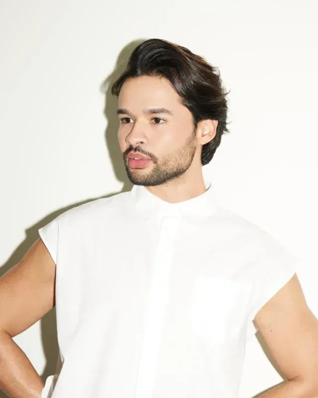 Man with dark hair and beard, wearing a white sleeveless shirt, looking to the side.