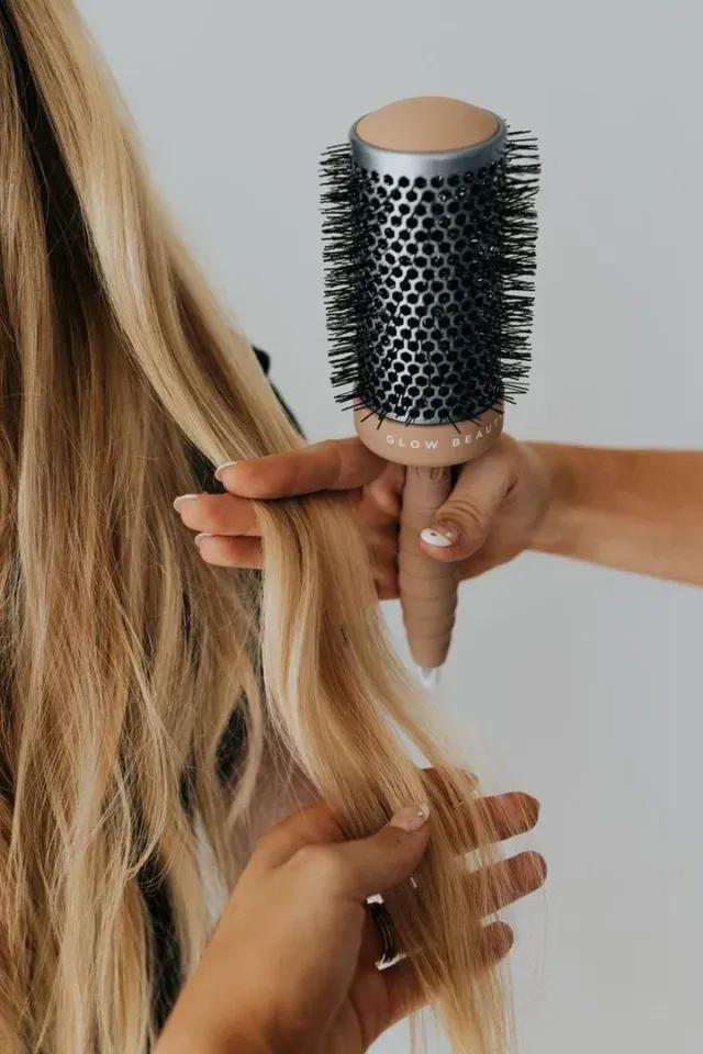 Woman styling long blonde hair with a round brush against a white background.