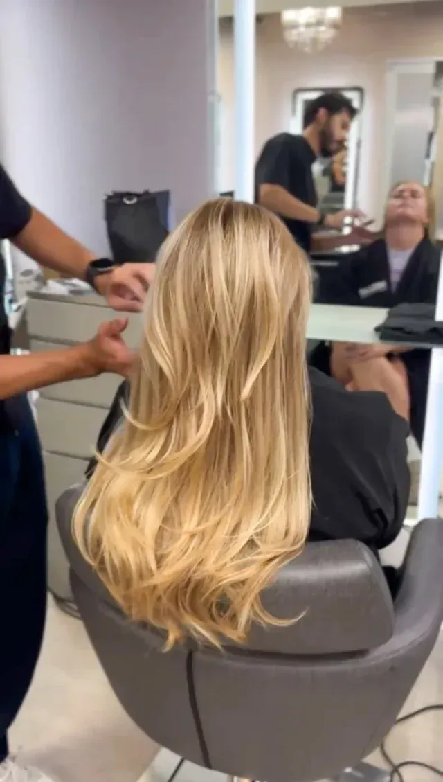 Person with long blonde hair in a salon chair, hairstylist working on it. Another person in background.
