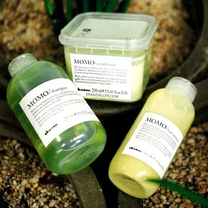 Three Davines MOMO hair care products: shampoo, conditioner, and hair mask, on a surface with pebbles and greenery.