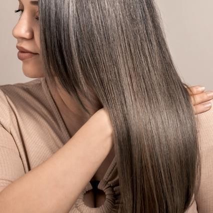 Woman with straight, gray hair, touching it with her hand. Light beige top.