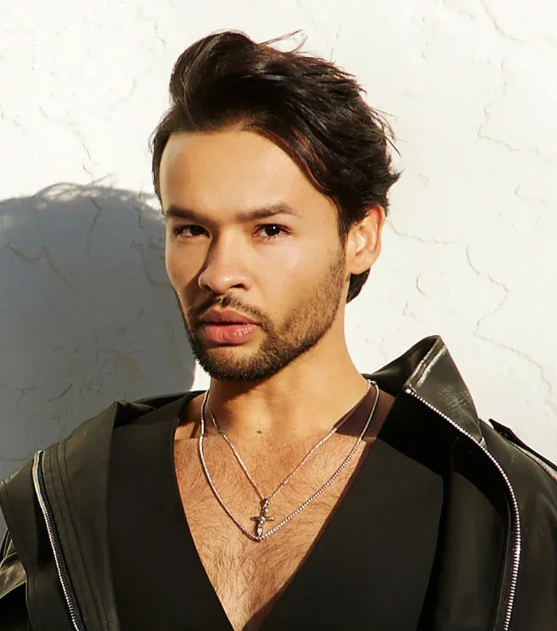 Man with dark hair and stubble in black outfit, looking toward camera.