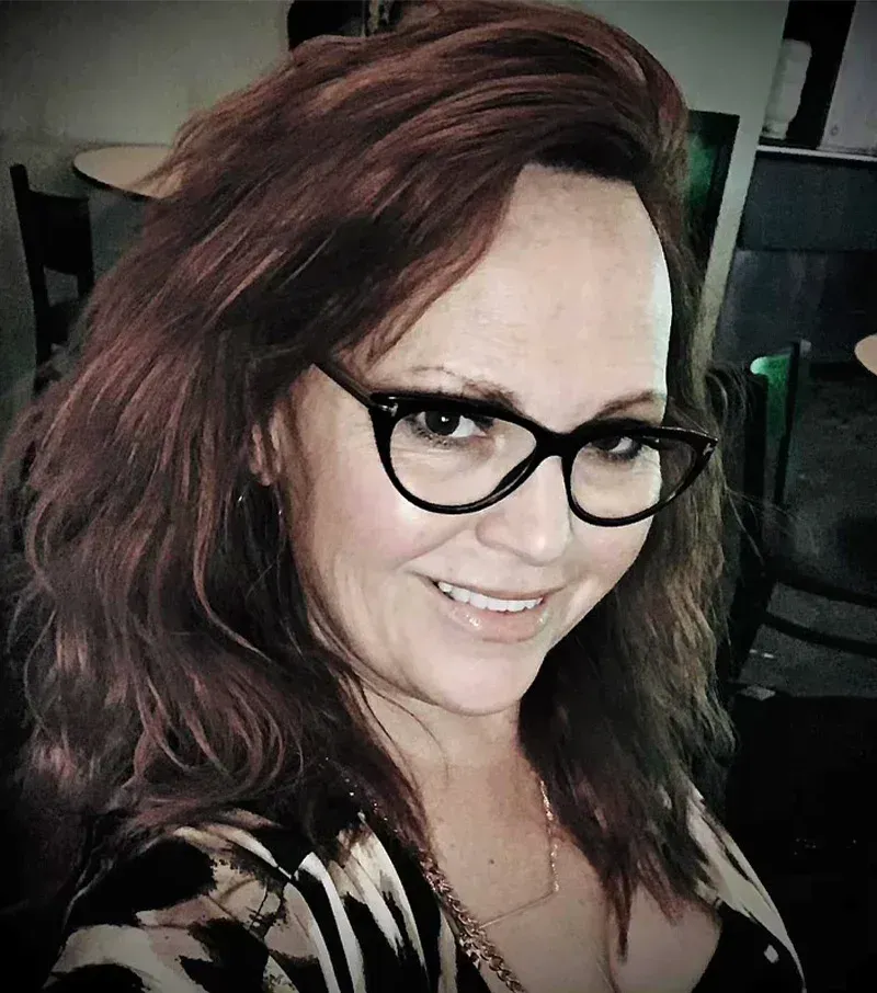 Woman with brown hair and glasses smiles; in a dark setting, wearing a patterned top.