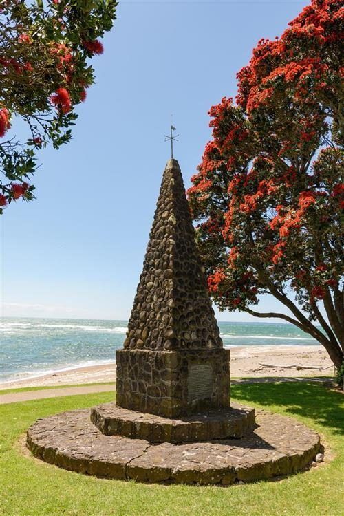 A stone obelisk with a cross on top of it is in a park next to a tree.