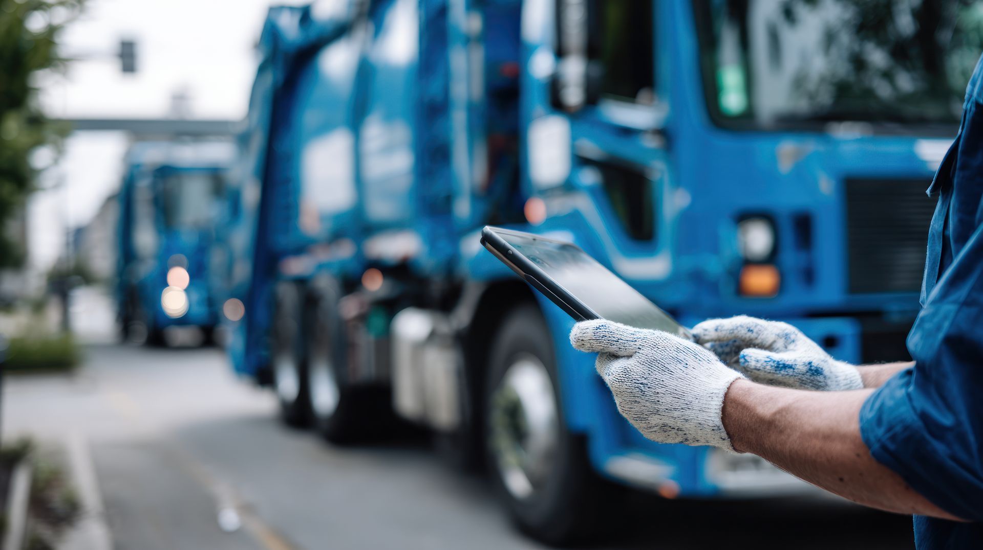 Worker uses tablet near blue sanitation truck on street. 