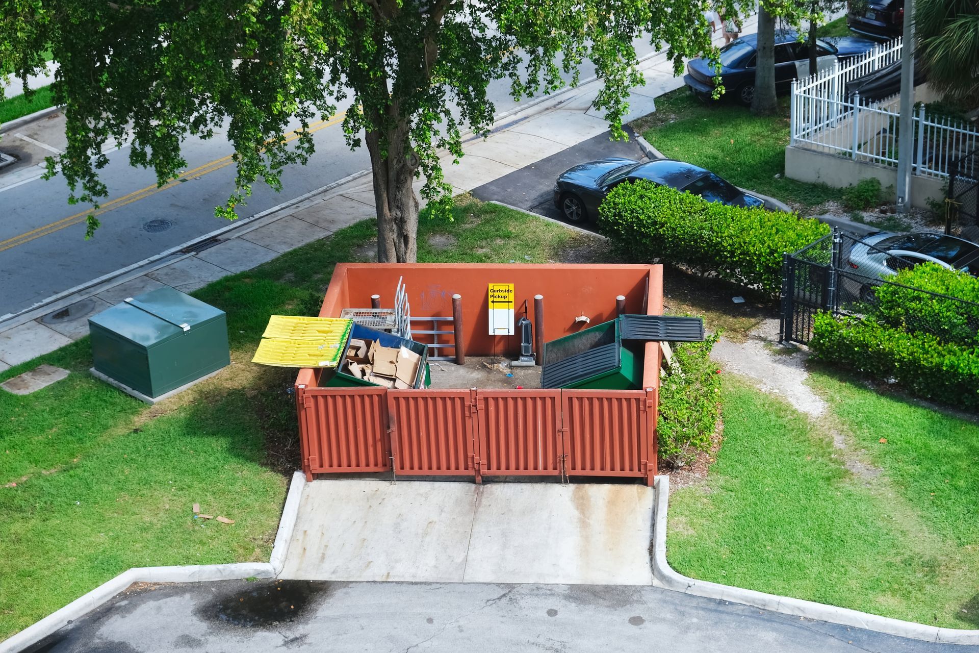 Aerial view of fenced garbage area with discarded items and nearby tree. Aerial view of fenced garbage area with discarded items and nearby tree.