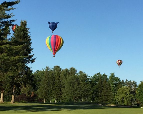 Four hot air balloons float over a tree-lined field against a clear blue sky.