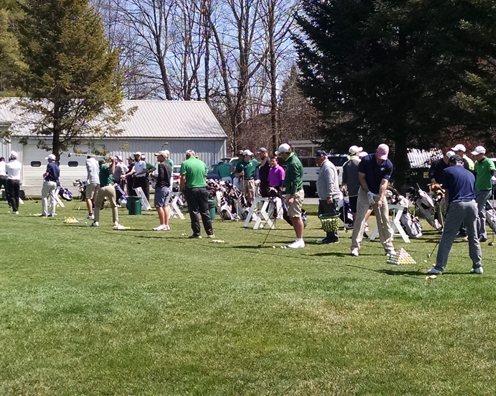 A group of people wearing casual clothing and hats practice swinging golf clubs on a sunny outdoor grass driving range.