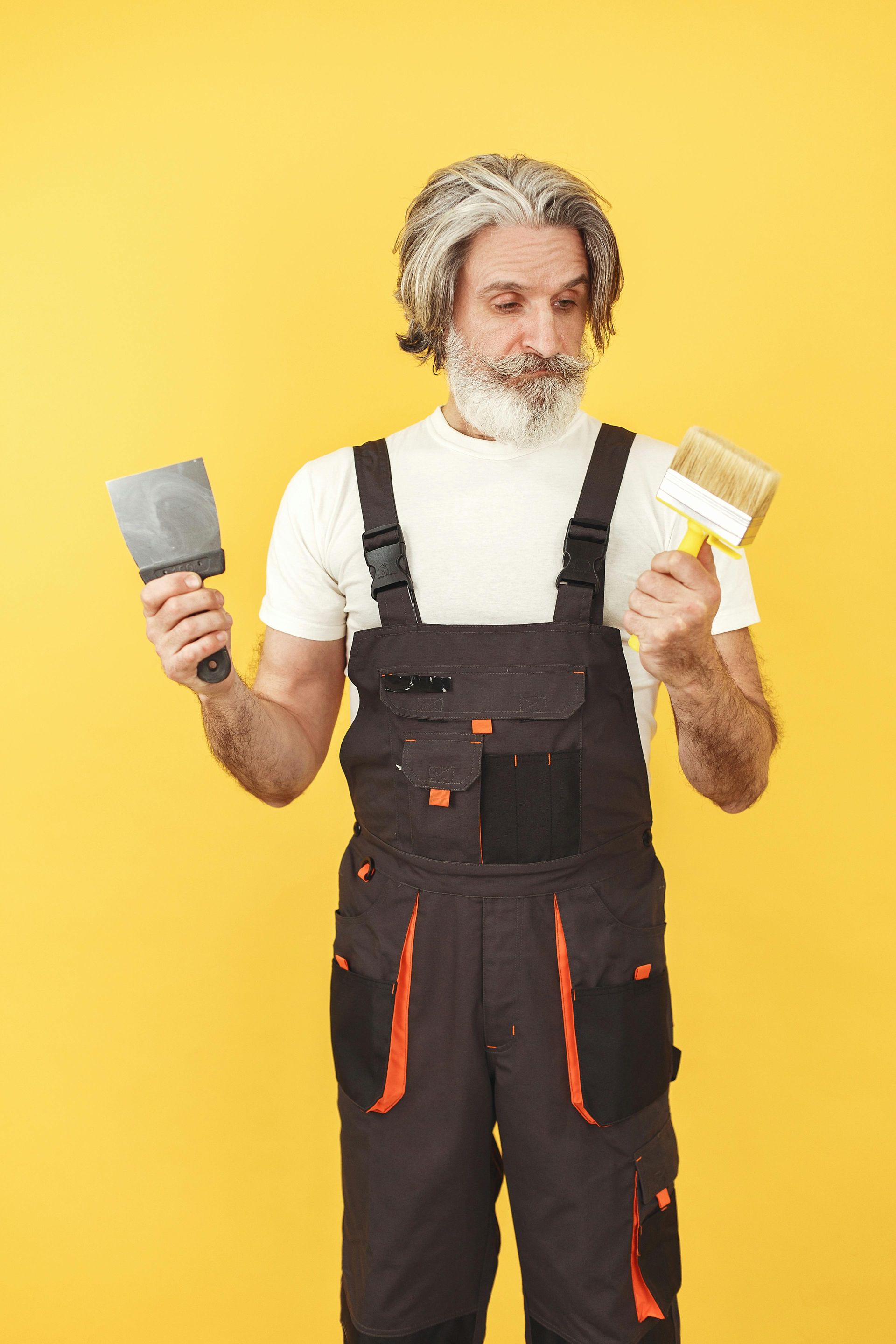 A man in overalls is holding a spatula and a paint brush.