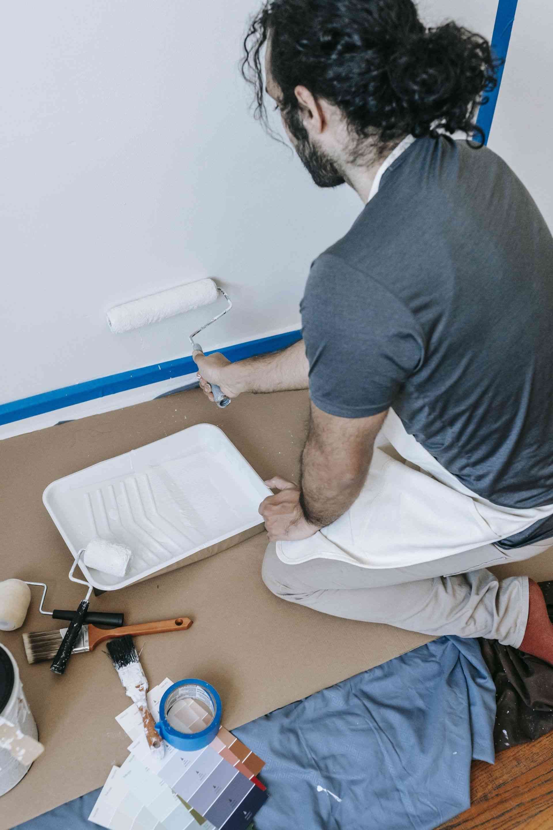 A man is kneeling down while painting a wall with a paint roller.