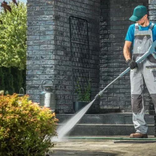 A man is using a high pressure washer to clean the sidewalk in front of a brick building.