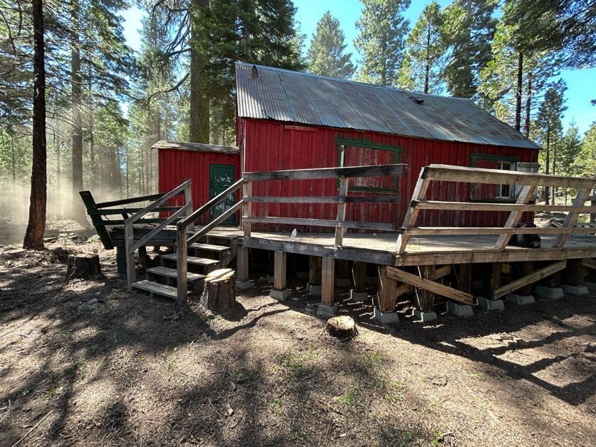 Red cabin with wooden deck in a forest. Sunlight filters through trees.