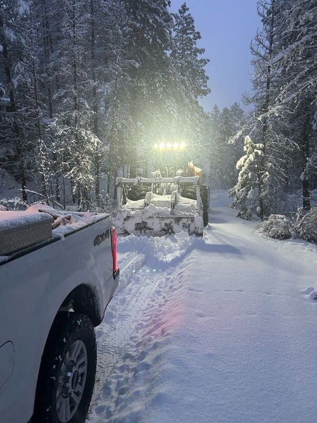 White pickup truck following a snowplow clearing a snow-covered road in a forest.