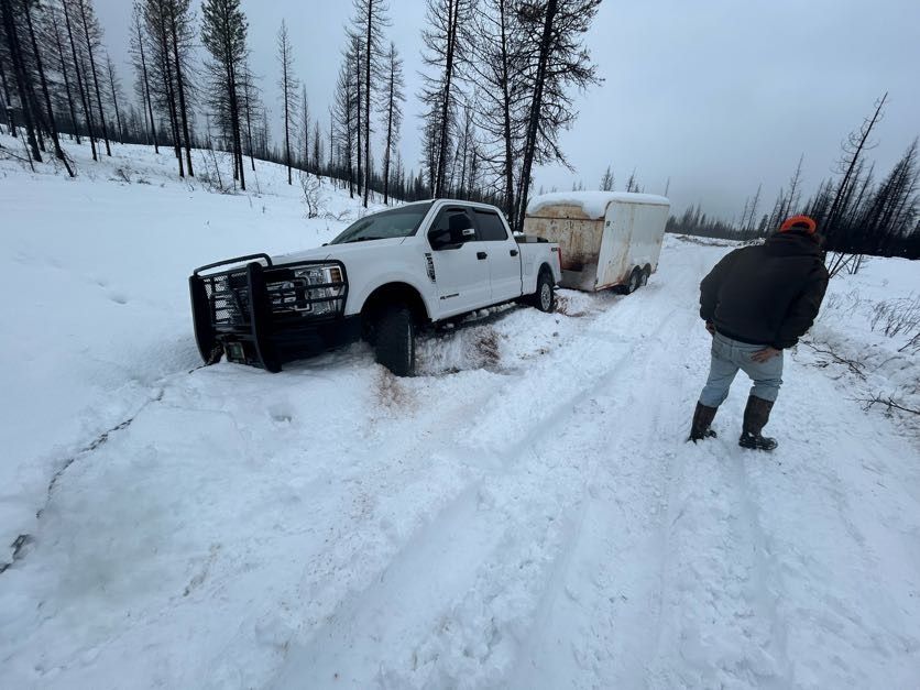 White truck and trailer stuck in snowy road, man inspecting. Forest backdrop.