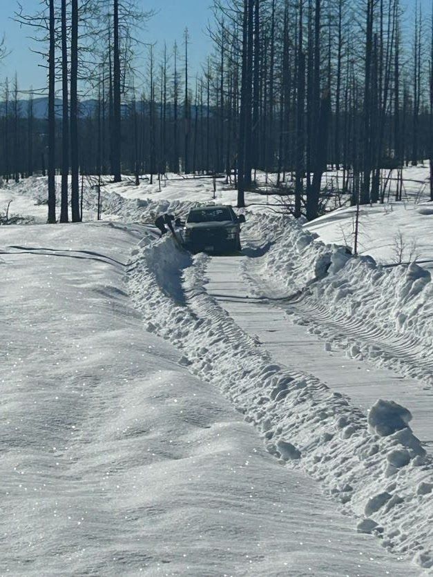 A vehicle travels down a snow-cleared road flanked by high walls of snow in a snowy forest.