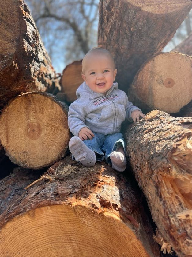Smiling baby sitting on logs. Wearing gray hoodie and blue jeans.