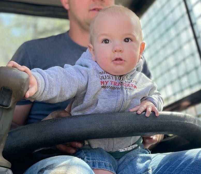 Baby sitting on a steering wheel with a man in a tractor; light-skinned with a gray hoodie, looking up.
