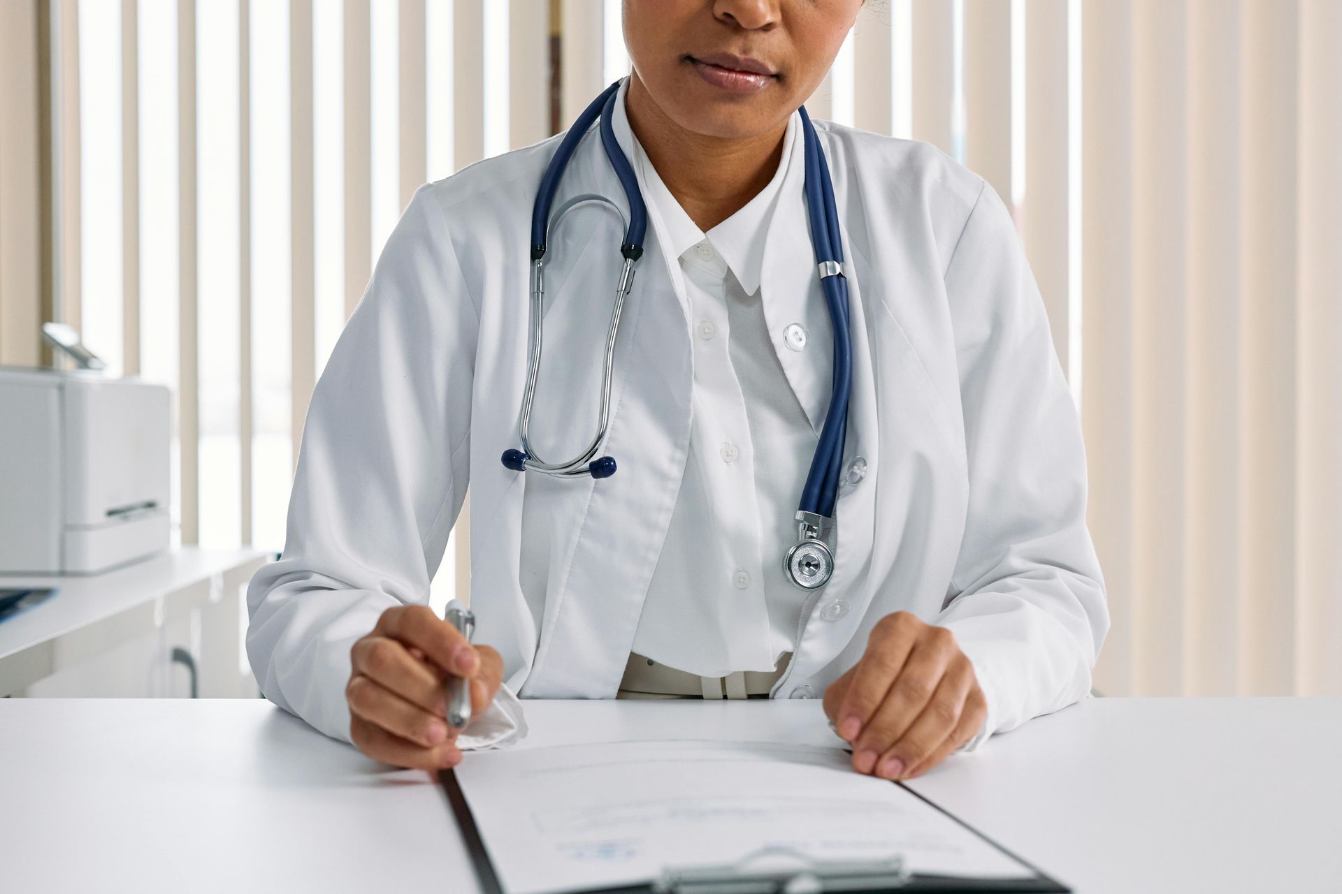 A female doctor is sitting at a desk writing on a clipboard.