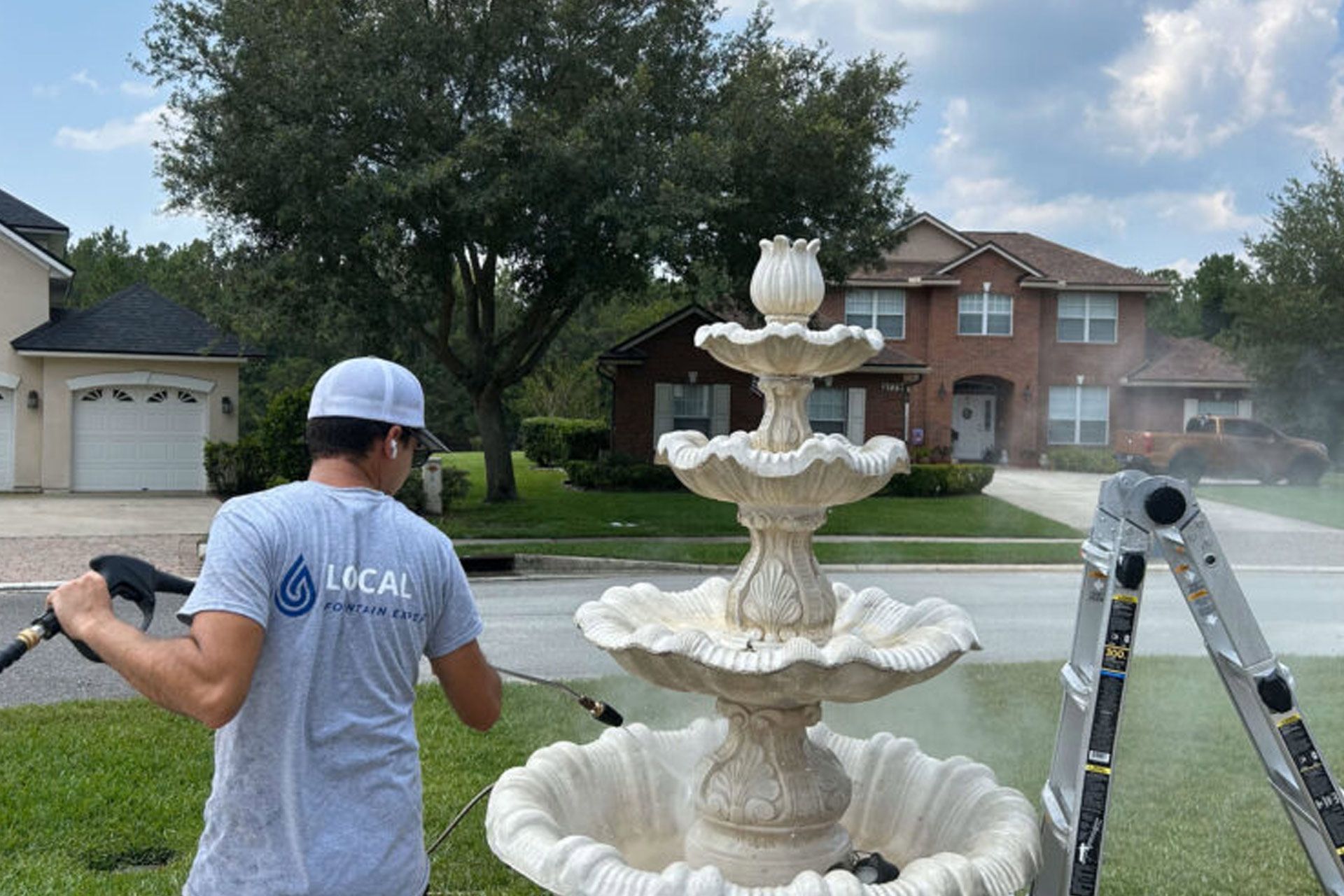 Man pressure washing a tiered outdoor fountain on a residential lawn. Houses and street in background.