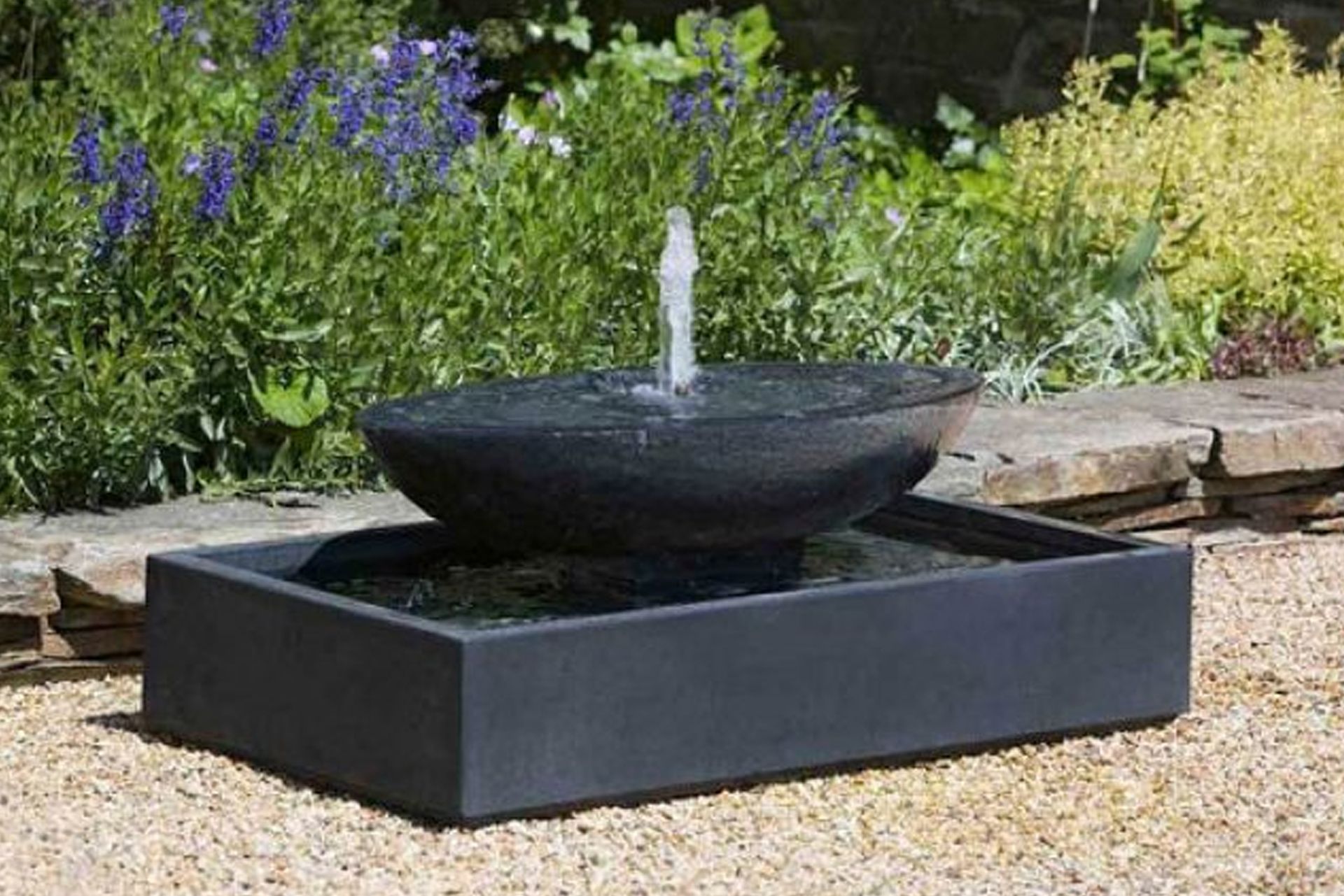 Circular flower bed with shrubs in front of a house, person working on the water fountain in the background.