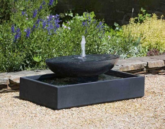 Circular flower bed with shrubs in front of a house, person working on the water fountain in the background.