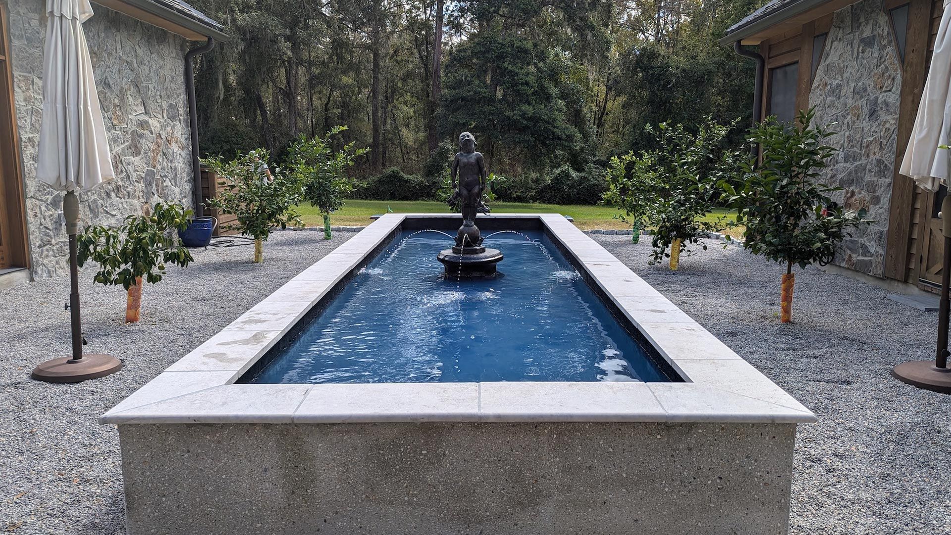 Rectangular water fountain with a statue, surrounded by trees and gravel patio.