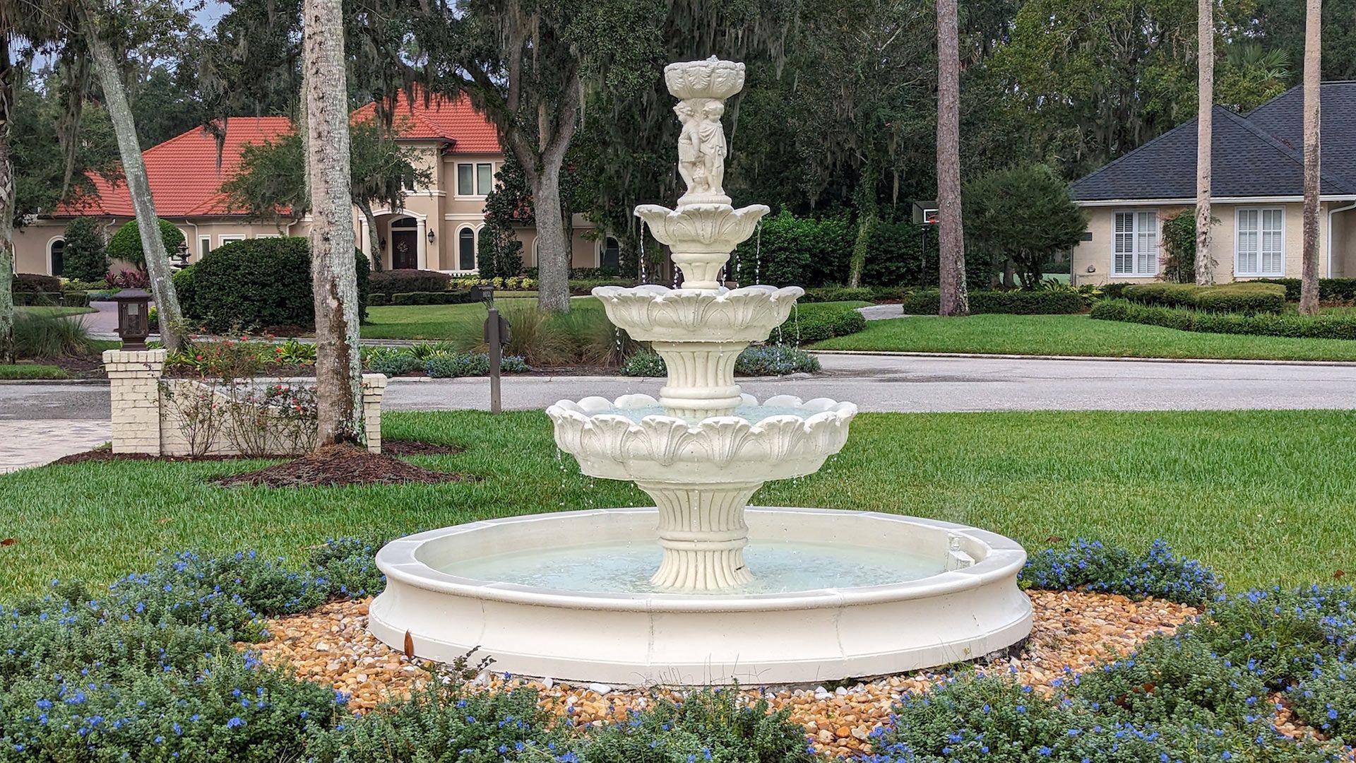 Three-tiered white fountain in a landscaped yard. Houses with red and black roofs are visible in the background.