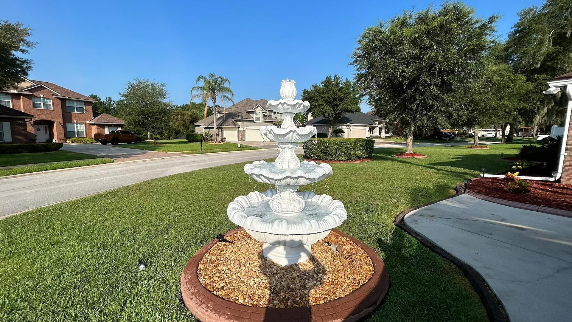 White tiered fountain in a grassy yard, surrounded by brown stones, with homes and trees in the background under a blue sky.