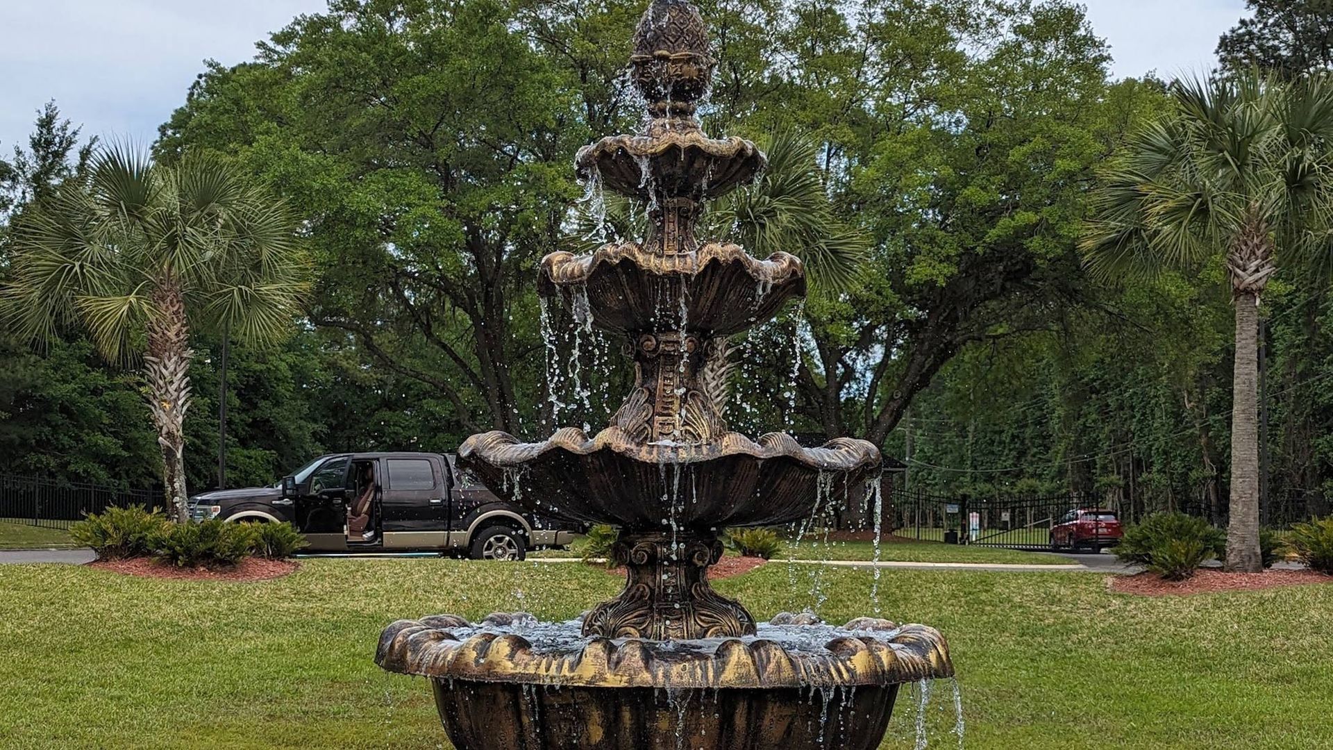 Tiered fountain with water cascading, in a grassy area with trees and a parked truck.