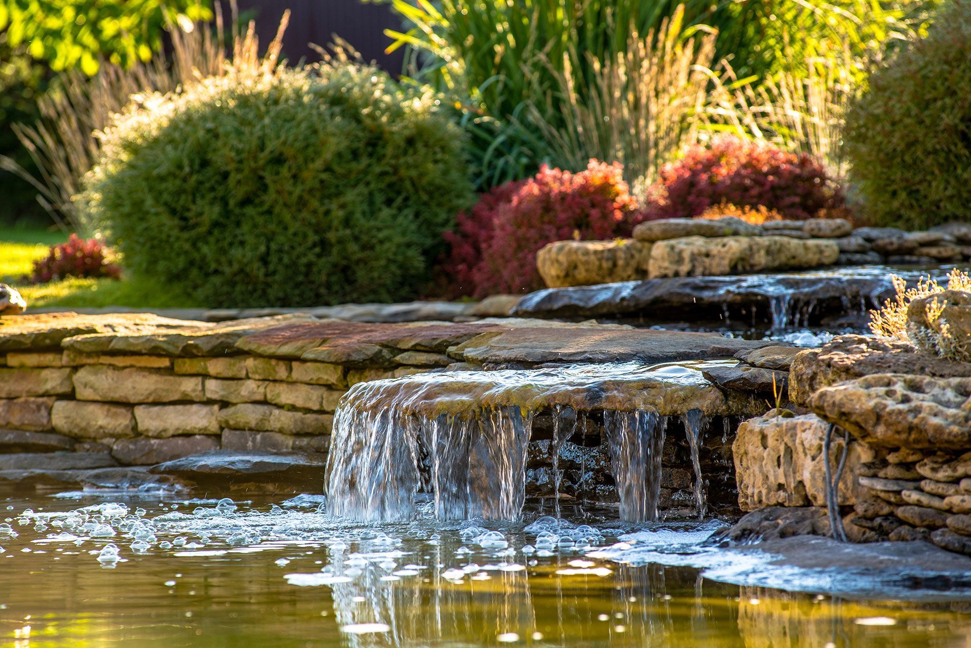 Water cascading over stone tiers into a pond, surrounded by lush greenery and sunlight.